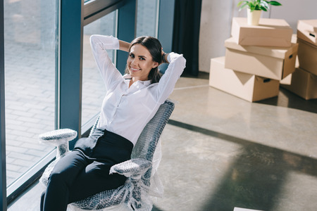 high angle view of happy young businesswoman sitting with hands behind head and smiling at camera in new officeの写真素材