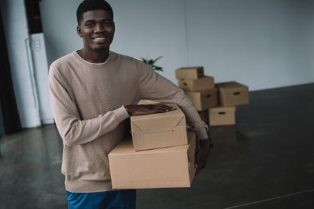 young african american man holding boxes and smiling at camera while relocating in new office