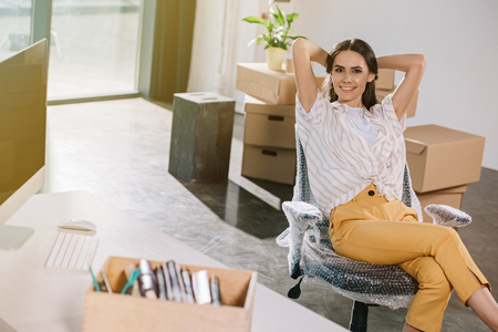happy young woman sitting with hands behind head and smiling at camera in new officeの写真素材