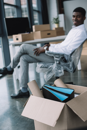 close-up view of cardboard box with office supplies and african american businessman sitting behind in new officeの写真素材