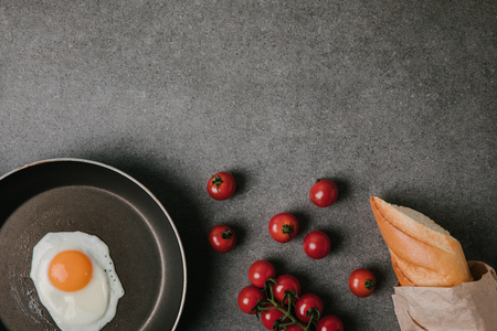 top view of fried egg on frying pan, fresh tomatoes and baguette in paper bag on greyの写真素材