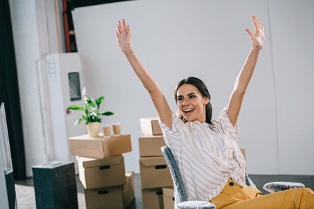 excited young businesswoman raising hands and looking away in new officeの写真素材