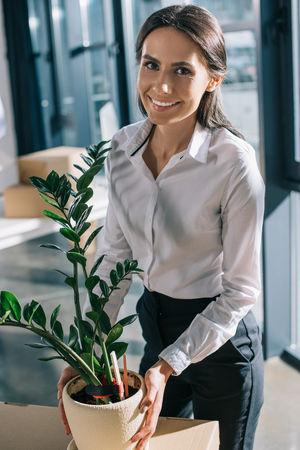 young businesswoman holding potted plant and smiling at camera in new officeの写真素材
