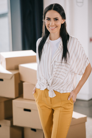 woman standing with hands in pockets and smiling at camera while relocating in new officeの写真素材