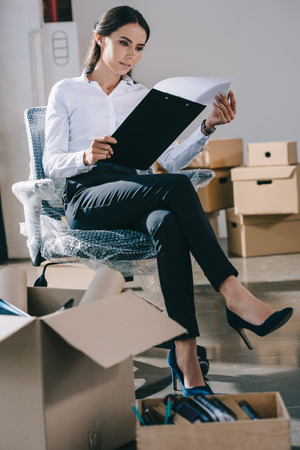 focused young businesswoman holding clipboard while sitting in new office during relocationの写真素材
