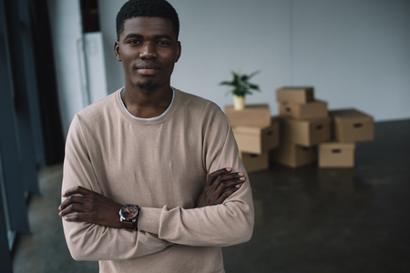 young african american man standing with crossed arms and looking at camera while relocating in new officeの写真素材