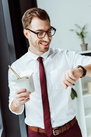 smiling businessman holding box with noodles and looking at watchの写真素材
