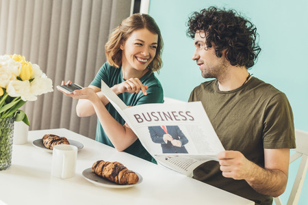 happy couple with smartphone and newspaper having breakfast together at homeの写真素材
