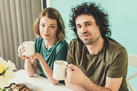 couple in t-shirts holding cups and looking at camera while having breakfast togetherの写真素材