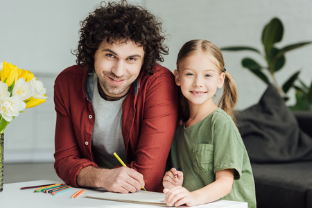 happy father and daughter drawing together and smiling at camera at homeの写真素材