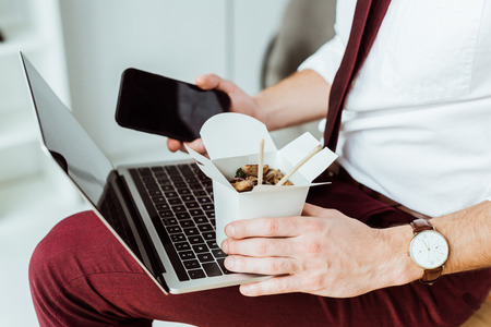 cropped view of businessman holding box with noodles, smartphone and laptopの写真素材