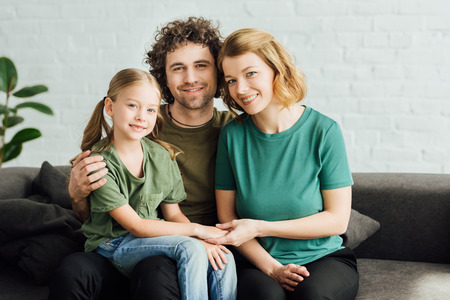 happy parents with cute little daughter sitting on couch and smiling at camera