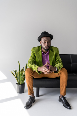 stylish african american in hat sitting on black sofa o grey backgroundの写真素材