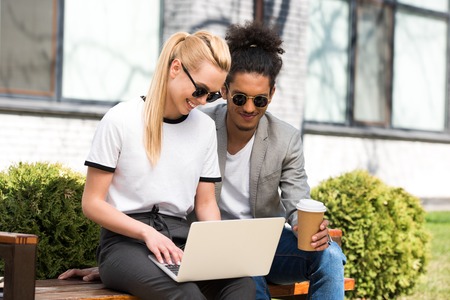 smiling young multiethnic couple using laptop and drinking coffee to go while sitting together on benchの写真素材