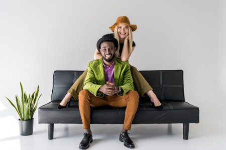 cheerful multicultural fashionable couple in hats sitting on black sofa on grey backgroundの写真素材