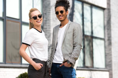 portrait of beautiful stylish multiethnic couple in sunglasses standing with hands in pockets and...