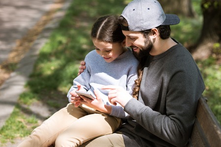happy father and daughter using smartphone on bench in parkの写真素材