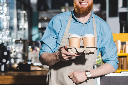 cropped image of smiling male barista holding two disposable cups of coffeeの写真素材