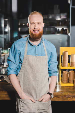 smiling young male barista in apron standing in coffee shopの写真素材