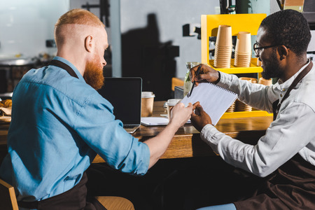 young african american male barista showing textbook to colleague in coffee shopの写真素材