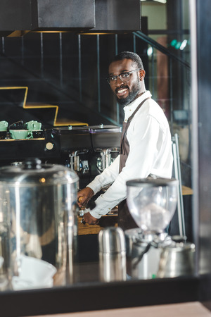 handsome young african american barista in eyeglasses smiling at camera while making coffee at coffee machineの写真素材
