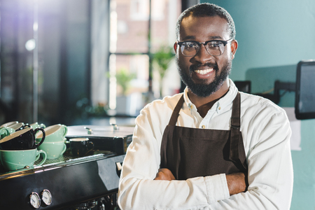 cheerful african american barista in apron and eyeglasses standing with crossed arms and smiling at camera in cafeの写真素材