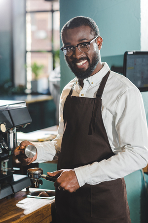 young african american barista in eyeglasses smiling at camera while making coffee at coffee machineの写真素材
