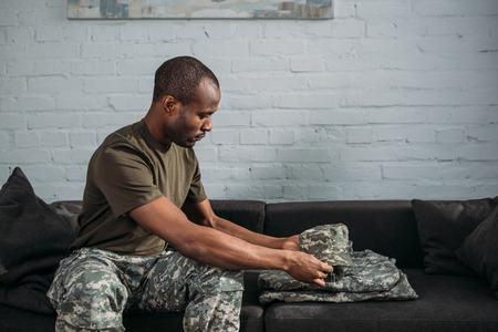African american male soldier folding camouflage clothes on sofaの写真素材