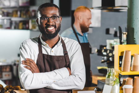 portrait of young smiling african american male barista in apron with colleague standing behindの写真素材