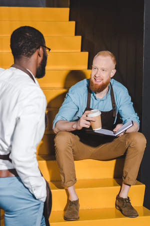 two multiethnic male baristas looking at each other and discussing work on stairsの写真素材