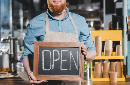 cropped shot of young male barista holding chalkboard with lettering open in coffee shopの写真素材