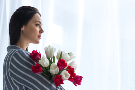 side view of beautiful pensive woman with bouquet of tulips at homeの写真素材