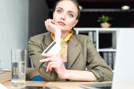 pensive stylish businesswoman holding credit card while sitting at workplaceの写真素材