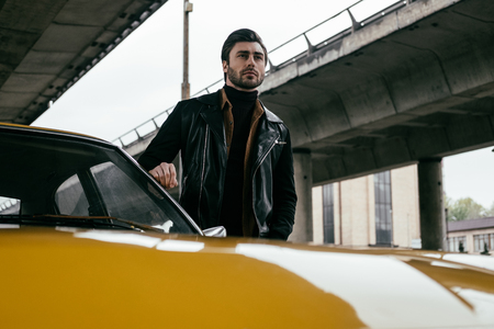 handsome stylish young man in leather jacket standing near yellow classic car and looking awayの写真素材
