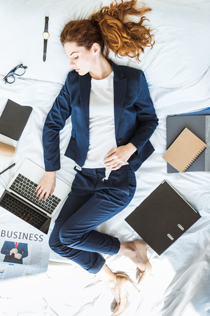Top view of businesswoman in suit typing on laptop while lying in bed among folders and documentsの写真素材