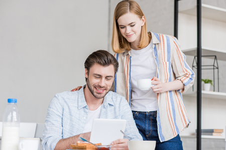 attractive woman holding boyfriend shoulder while he using digital tablet at table with breakfastの写真素材