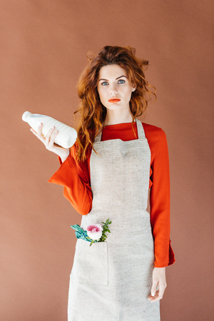 Woman with long red hair in apron holding milk bottle isolated on brown backgroundの写真素材