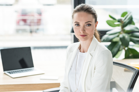 portrait of smiling beautiful businesswoman in white suit at workplace with laptop in officeの写真素材