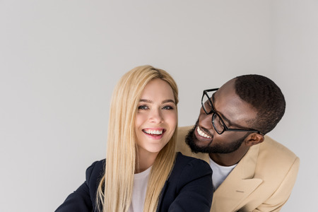 happy young african american man in eyeglasses lookign at beautiful blonde girlfriend isolated on greyの写真素材
