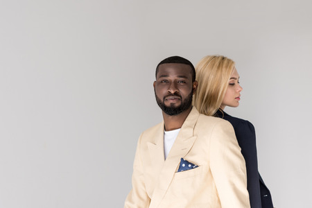 stylish young multiethnic couple sitting back to back, man looking at camera isolated on greyの写真素材