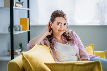 portrait of attractive woman sitting on sofa and looking at camera at homeの写真素材