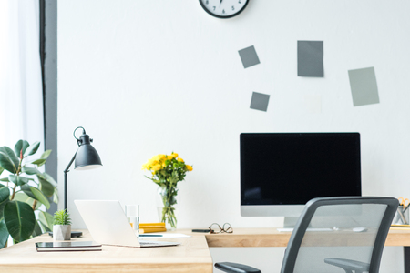 close up view of empty workplace with blank computer screen, laptop, eyeglasses and bouquet of chrysanthemum flowers in officeの写真素材