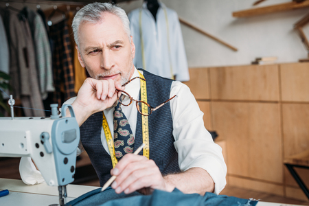 handsome pensive tailor sitting at table with sewing machine at sewing workshopの写真素材