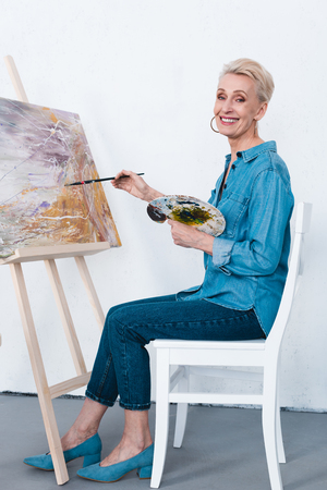 senior woman painting on easel with paintbrush and palette in workshopの写真素材