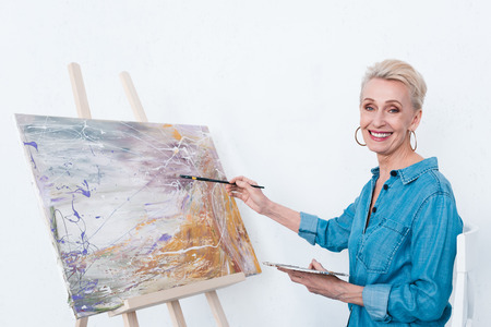 senior cheerful woman painting with palette on easel in workshopの写真素材