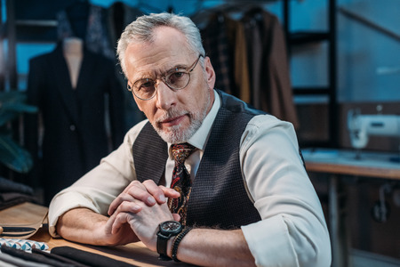 handsome mature tailor sitting at workplace at sewing workshop in late evening and looking at camera
