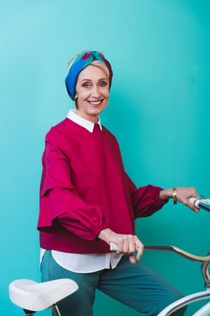 cheerful senior woman standing with bicycle, isolated on turquoiseの写真素材