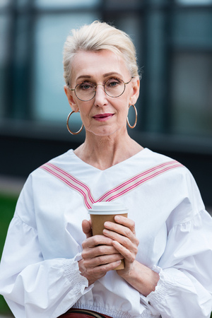 attractive stylish senior woman in trendy eyeglasses holding coffee to goの写真素材