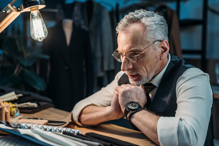 confident mature tailor looking at cloth samples at sewing workshop in late eveningの写真素材