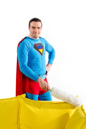 handsome male superhero cleaning couch with duster and smiling at camera isolated on whiteの写真素材